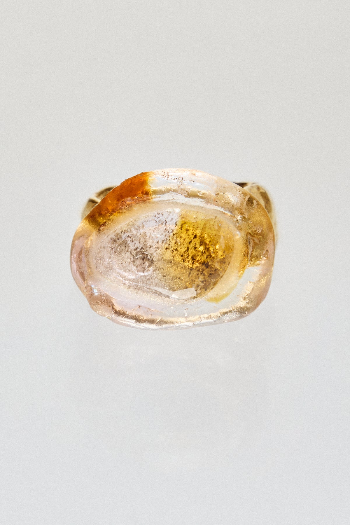 A unique ring featuring a large, organic-shaped clear resin cabochon filled with gold leaf and suspended amber-colored inclusions, set against a plain light gray background.