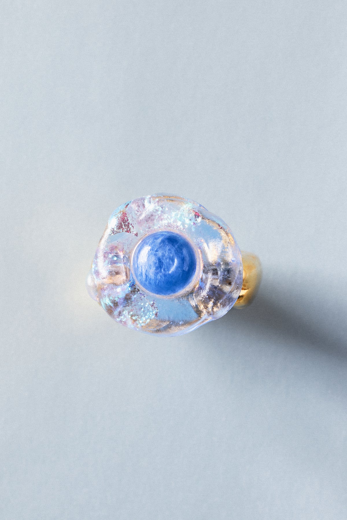 A handmade glass ring featuring a central round blue cabochon surrounded by a textured, iridescent clear glass halo with a gold band, shown on a neutral light gray background.