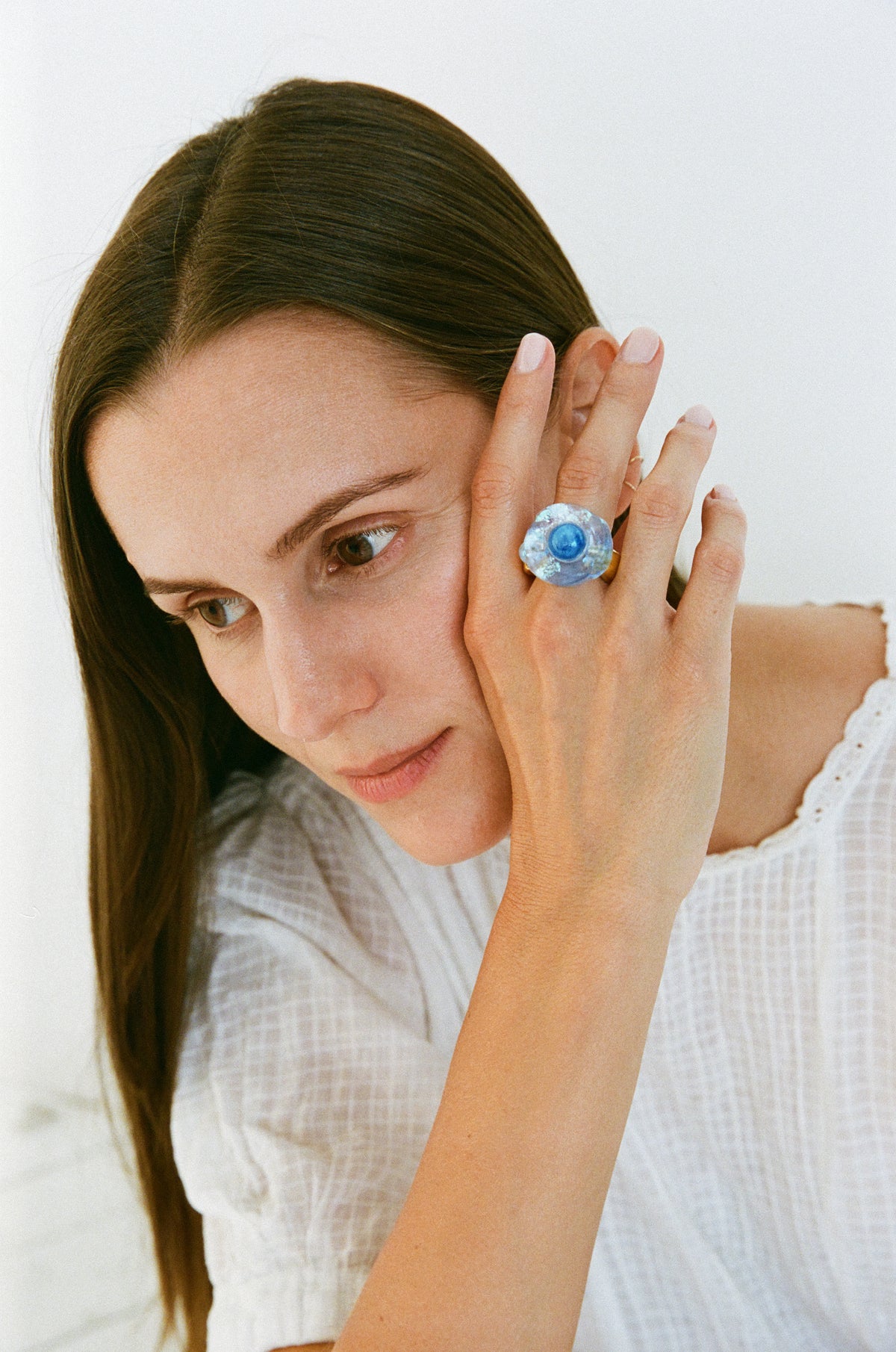 A woman with long brown hair wearing a white textured top, holding her hand near her ear to showcase a large, round blue and white evil eye ring.