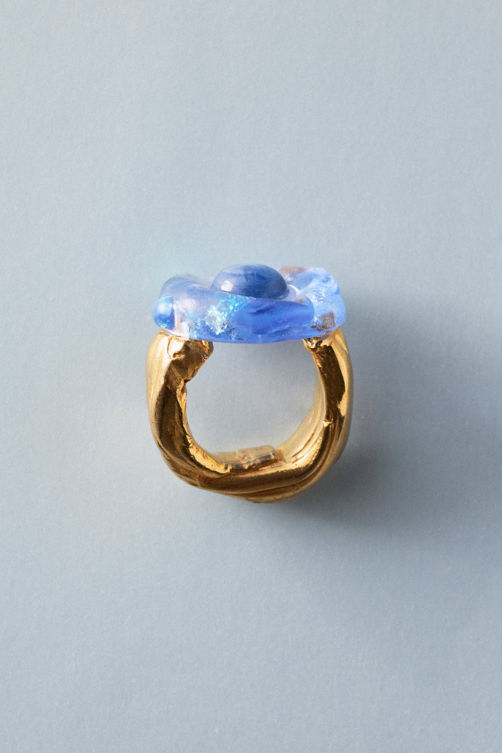 A gold ring with a sculptural, organic-shaped blue glass top, photographed on a solid light gray background.
