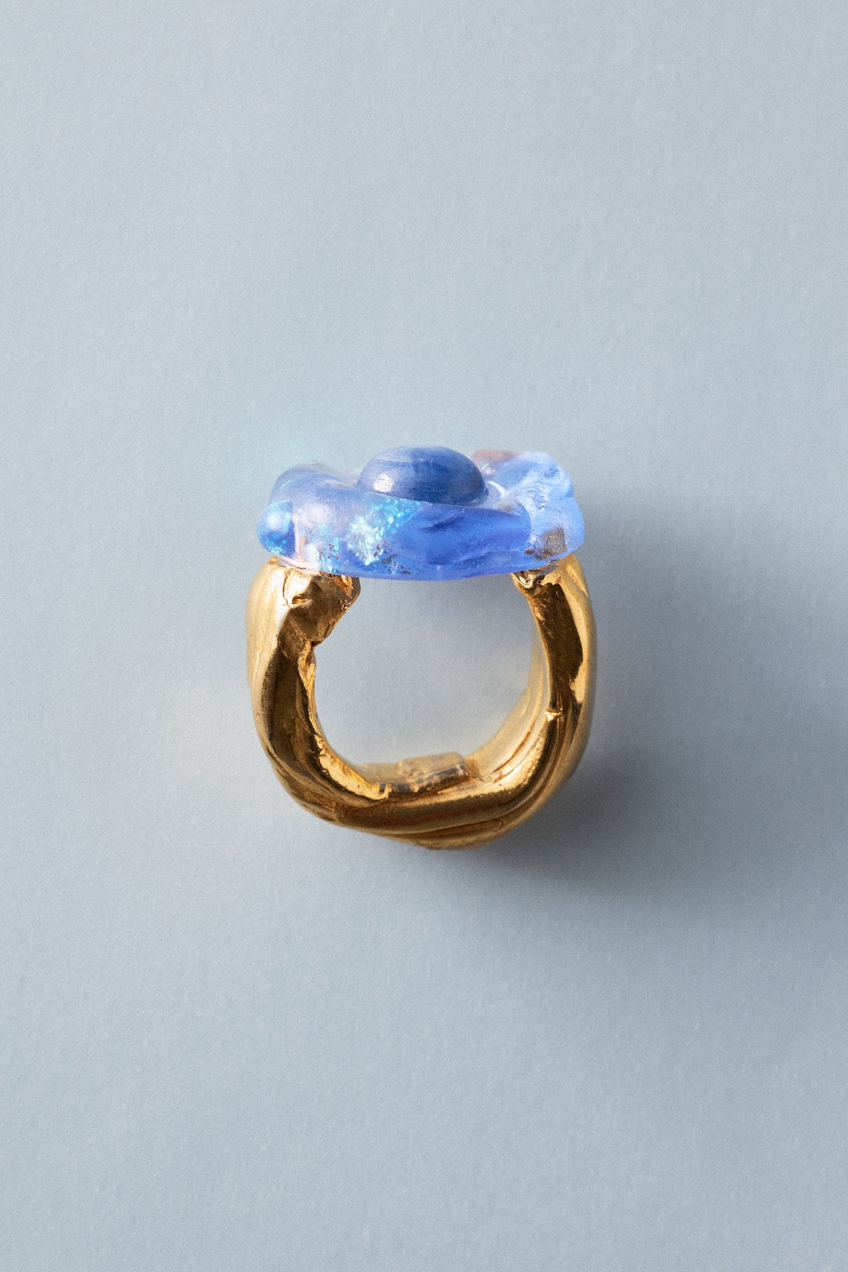 A gold ring with a sculptural, organic-shaped blue glass top, photographed on a solid light gray background.