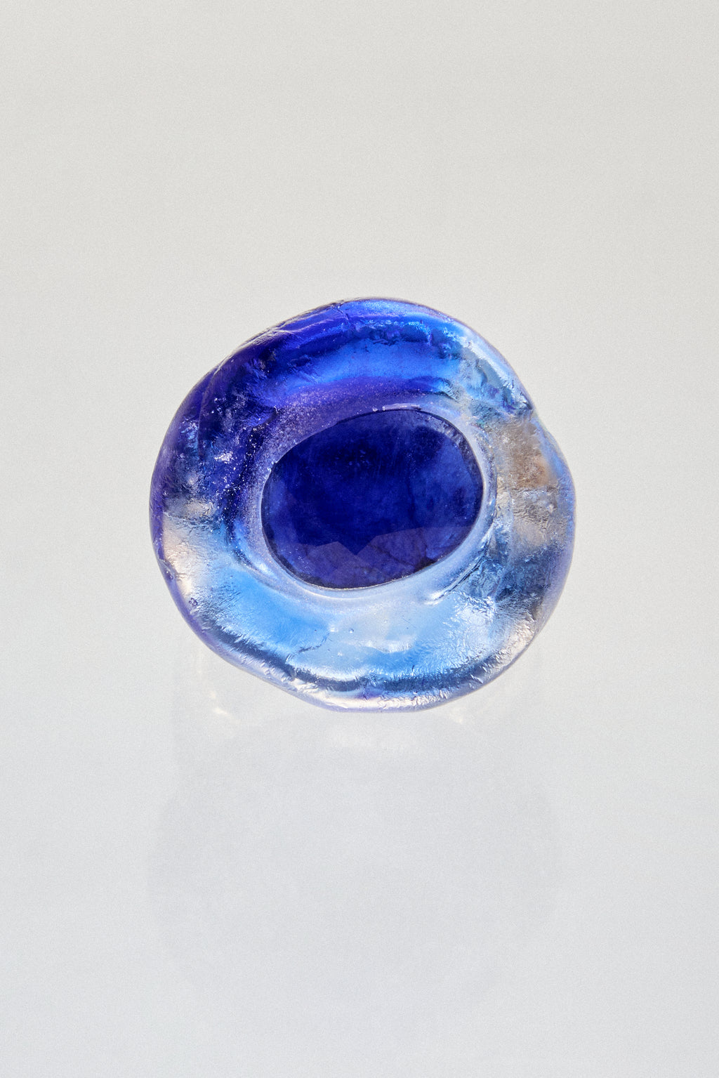 A handmade glass ring with a deep blue center and swirling light blue and white patterns, photographed on a clean white reflective surface.