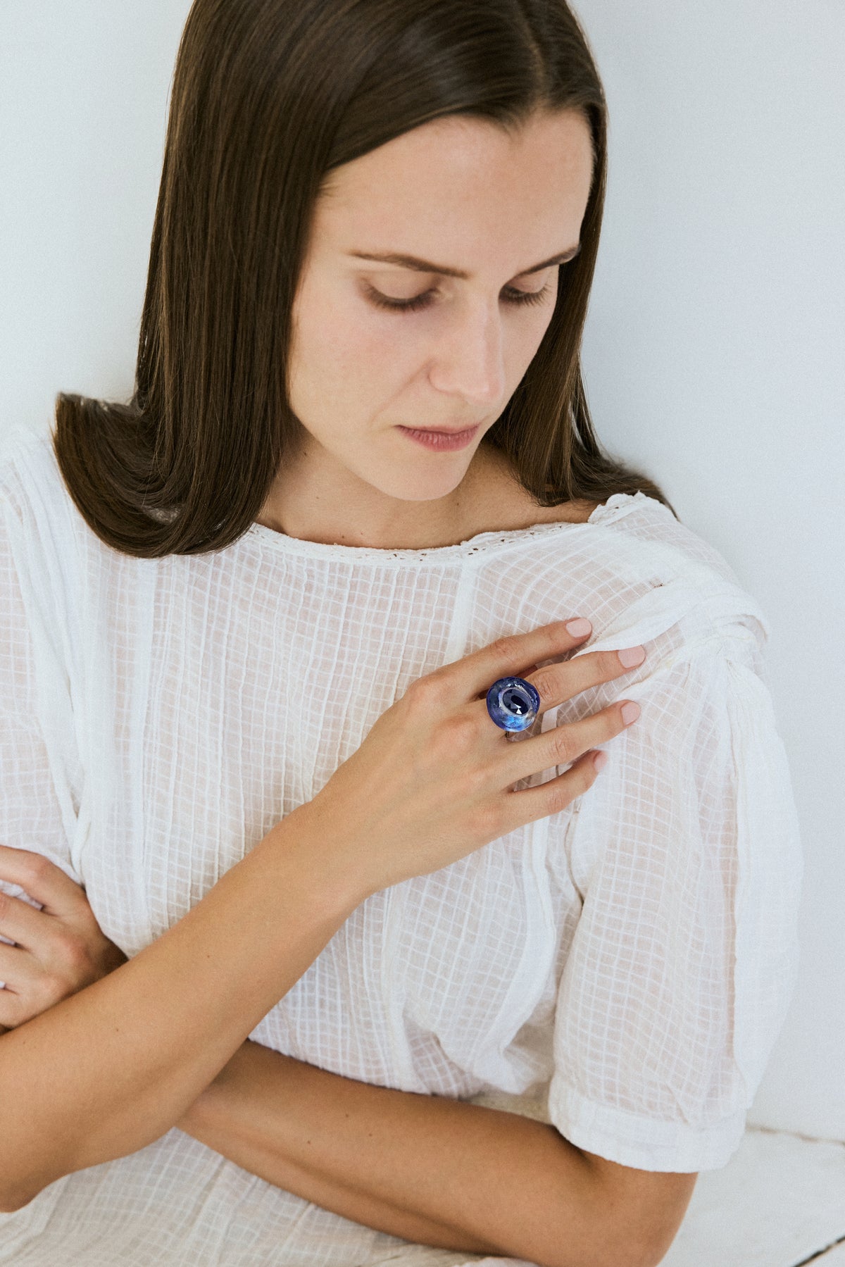 A woman wearing a white textured top and a large blue ring on her finger, looking down with a contemplative expression against a plain white background.