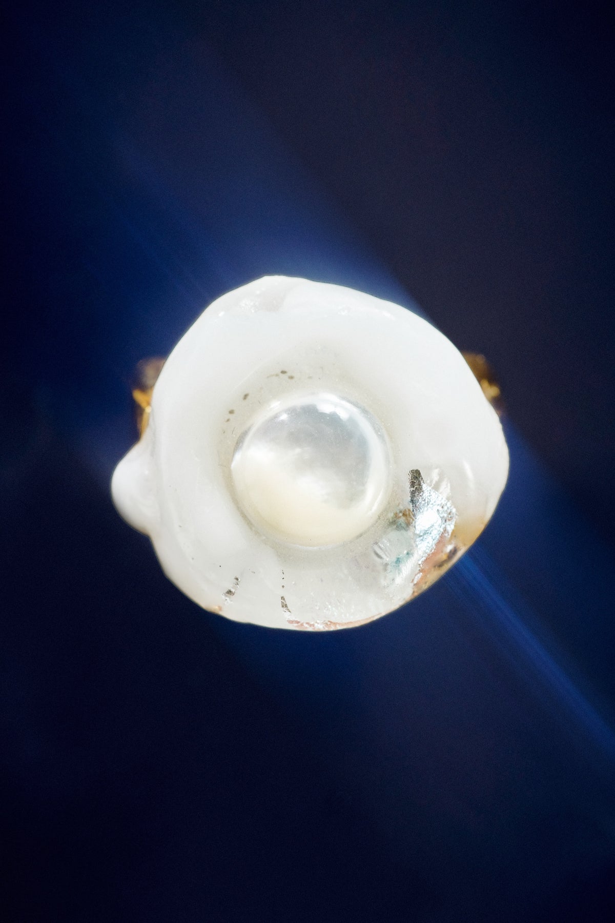 A close-up of a white irregularly shaped ring with a smooth, lustrous white pearl set in the center against a dark blue background.