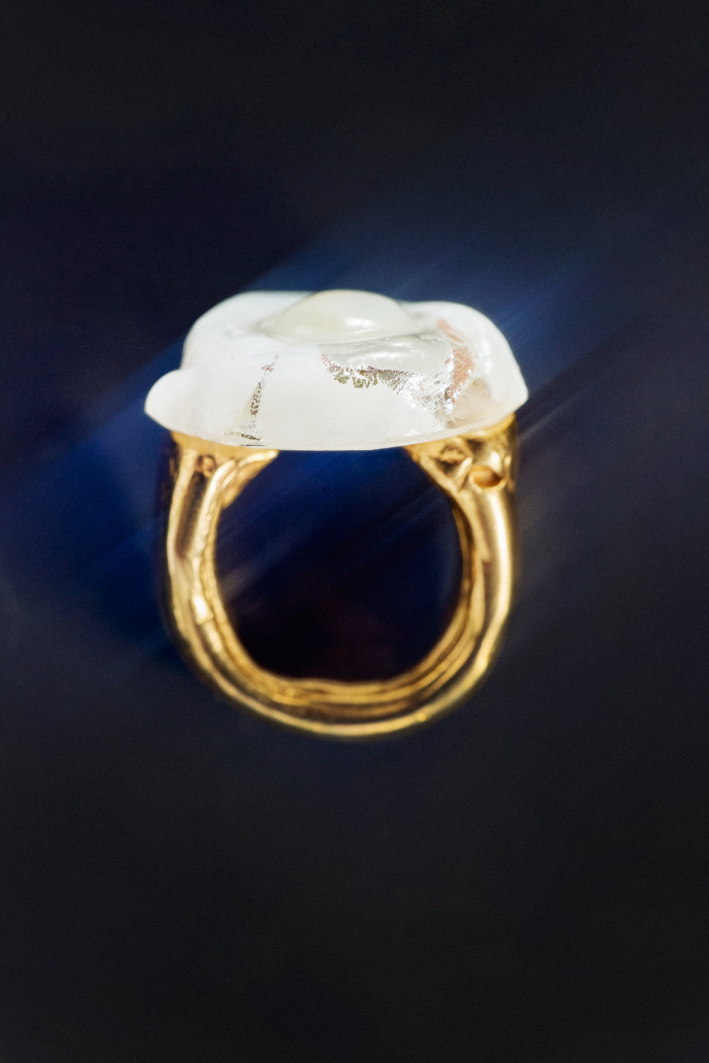 A gold ring with a large, irregularly shaped white stone featuring natural inclusions, set against a dark background with a blue light flare.