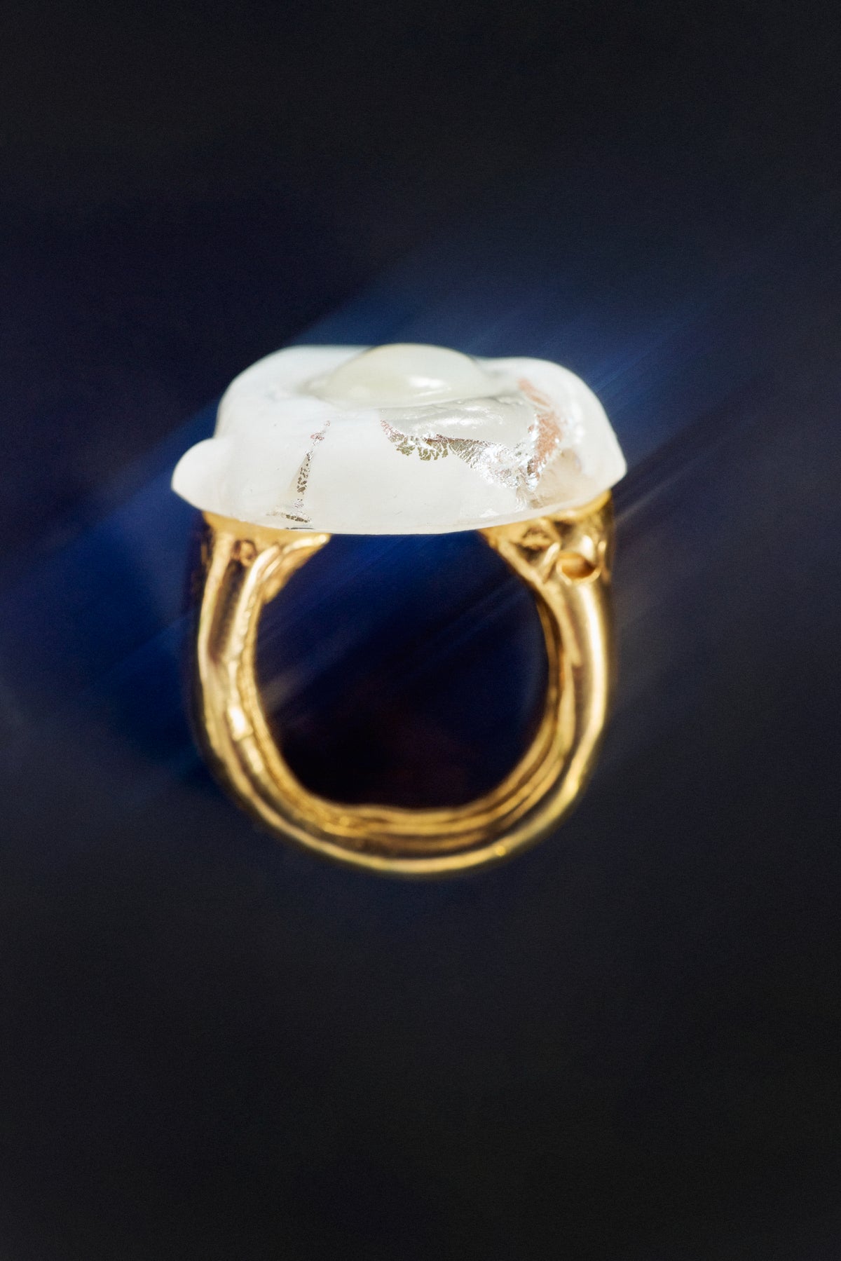 A gold ring with a large, irregularly shaped white stone featuring natural inclusions, set against a dark background with a blue light flare.